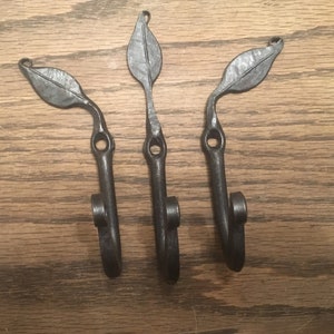 May include: Three handcrafted metal wall hooks. Each hook features a leaf-shaped top, a curved stem, and a hook at the bottom. The hooks are dark gray and appear to be made of iron or a similar metal. They are designed to be mounted on a wall.