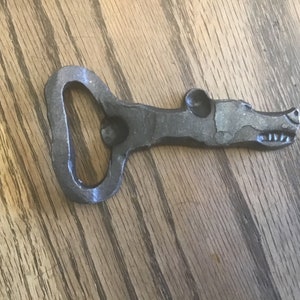 May include: A metal bottle opener shaped like an alligator's head. The opener is made of dark gray metal and has a detailed design.