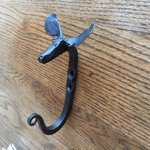 May include: Black metal moose head wall hook with a curved hook at the bottom. The hook is mounted on a wooden surface.
