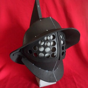 May include: A black gladiator helmet with a pointed crest and a face mask with a grid pattern. The helmet is made of foam and is sitting on a white base.