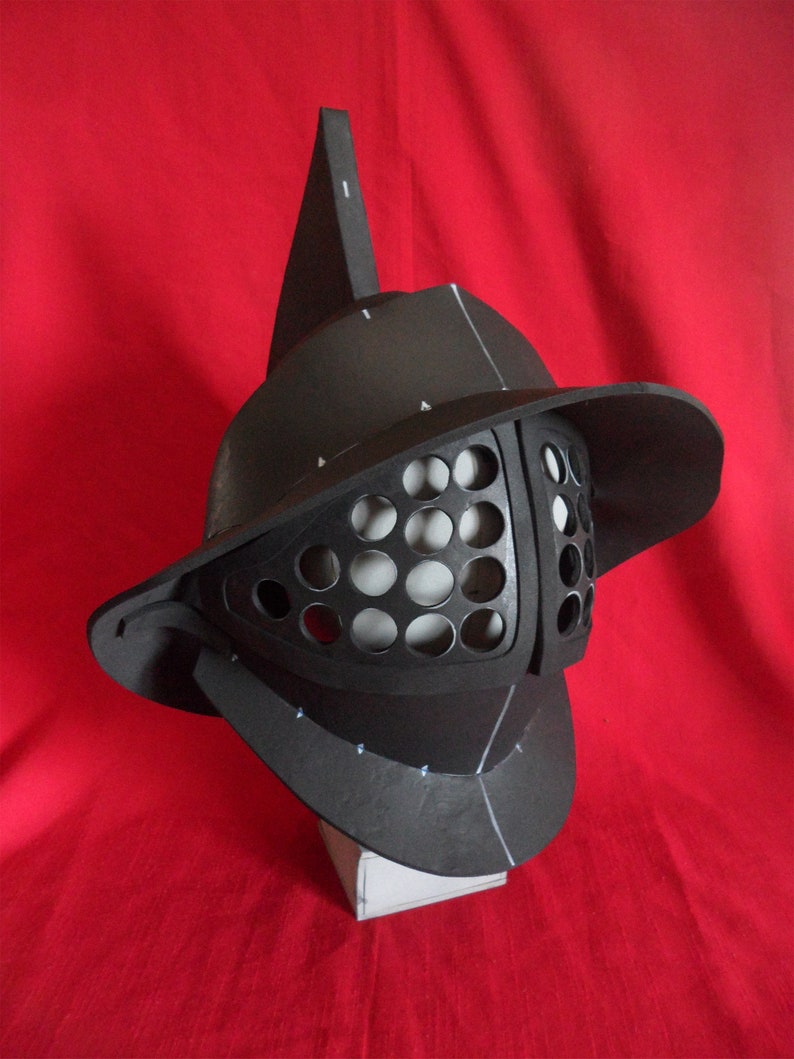 May include: A black gladiator helmet with a pointed crest and a face mask with a grid pattern. The helmet is made of foam and is sitting on a white base.
