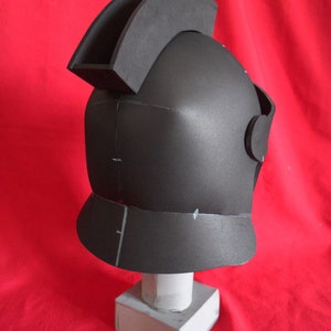 May include: A black foam helmet with a raised crest on top. The helmet is sitting on a white pedestal.