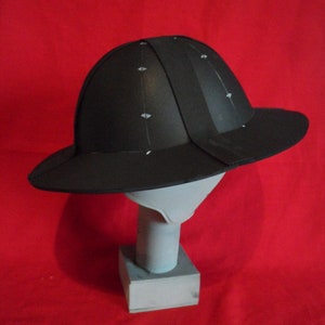 K&ouml;nnte beinhalten: Ein schwarzer Helm mit breitem Rand, auf einem wei&szlig;en St&auml;nder vor rotem Hintergrund.