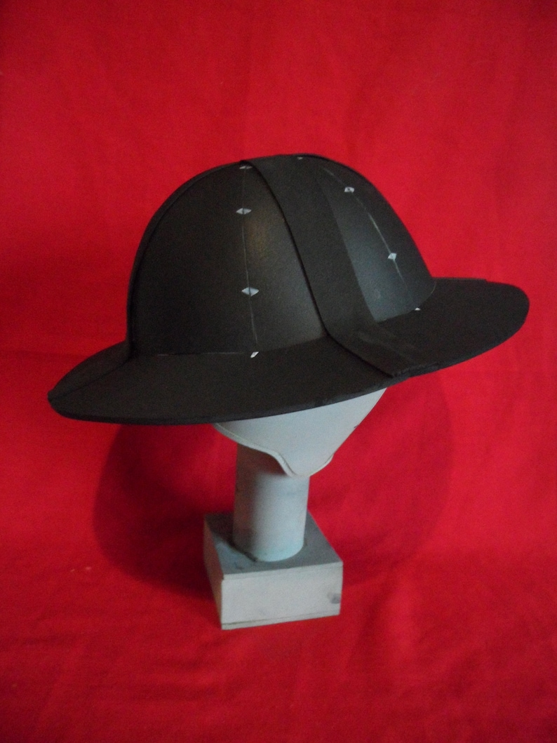 K&ouml;nnte beinhalten: Ein schwarzer Helm mit breitem Rand, auf einem wei&szlig;en St&auml;nder vor rotem Hintergrund.