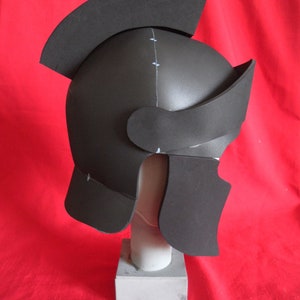 May include: A black foam helmet with a raised crest, resembling a Roman or Greek soldier's helmet. The helmet is on a white foam stand.