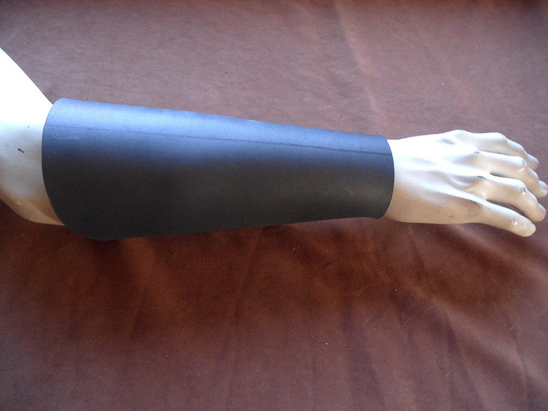 May include: A black arm guard made of a flexible material, covering the forearm. The arm guard is shown on a mannequin arm, with the hand visible. The guard appears to be a single piece, wrapping around the arm.