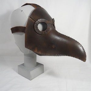 May include: A brown leather plague doctor mask with a long beak and eye holes. The mask is on a white mannequin head.