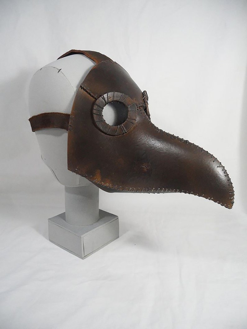 May include: A brown leather plague doctor mask with a long beak and eye holes. The mask is on a white mannequin head.