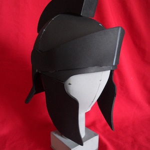 May include: A black foam helmet with a raised crest and a pointed chin guard. The helmet is on a white foam stand.