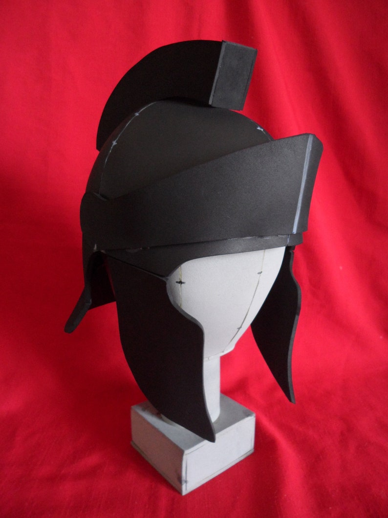 May include: A black foam helmet with a raised crest and a pointed chin guard. The helmet is on a white foam stand.
