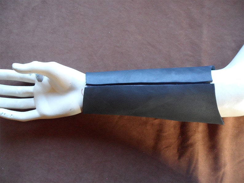 May include: Black leather arm cuff, shown on a mannequin hand. The cuff is made of two overlapping pieces of leather, with a curved edge. The background is a solid brown color.