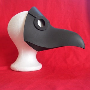 May include: A black plague doctor mask with a long beak and a round eye hole. The mask is made of foam and is displayed on a white mannequin head.
