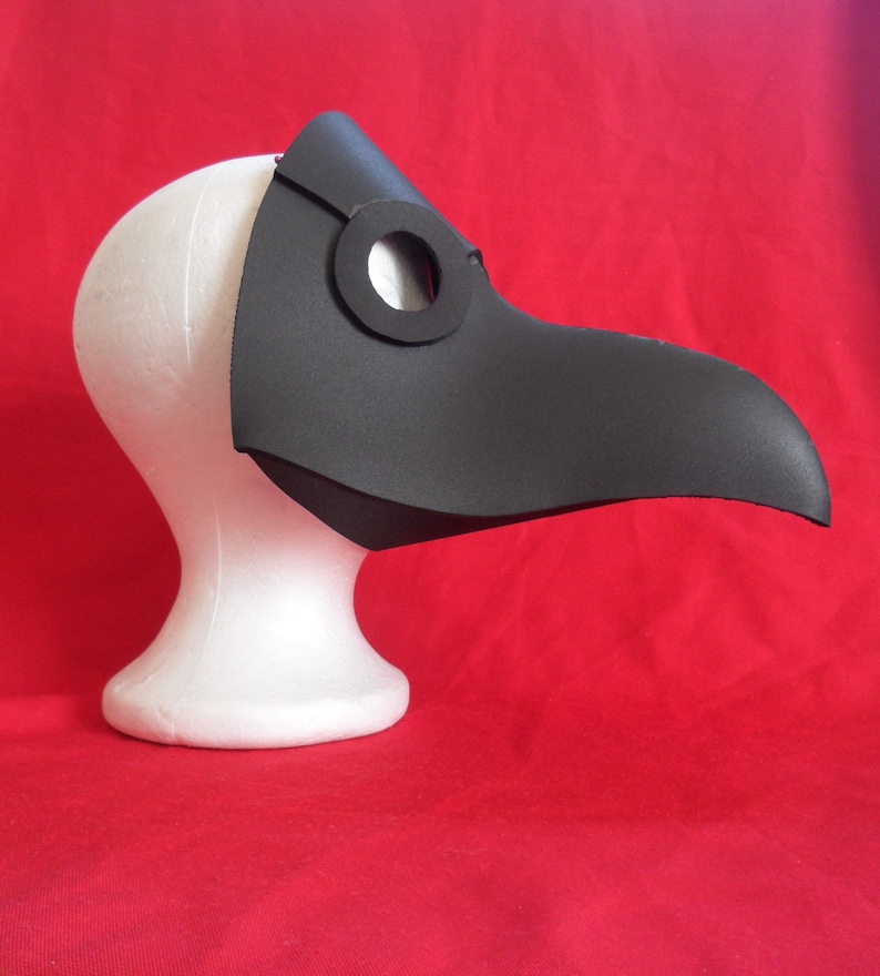 May include: A black plague doctor mask with a long beak and a round eye hole. The mask is made of foam and is displayed on a white mannequin head.
