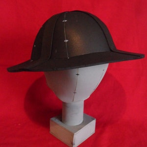 K&ouml;nnte beinhalten: Ein schwarzer Helm mit breitem Rand, der auf einem wei&szlig;en Mannequin-Kopf sitzt. Der Helm ist aus hartem Material gefertigt und hat eine glatte Oberfl&auml;che.