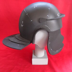 May include: A black foam helmet with a visor and a neck guard. The helmet is on a white stand and has markings on it.