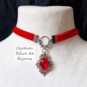 May include: A red velvet choker with a silver clasp and a pendant featuring a large red gemstone set in a silver filigree frame. The text "Cachou Rêve de Bijoux" is visible in the image.