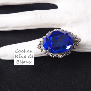 May include: A silver ring featuring a large, oval-shaped, blue gemstone set within an ornate, decorative frame. The ring is displayed on a white hand model.