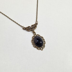 May include: A delicate gold necklace with a black oval gemstone pendant. The pendant is surrounded by an ornate, filigree design.