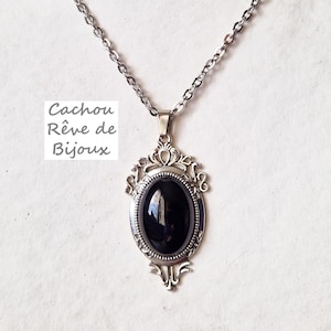 Op de afbeelding: Zilverkleurige hangerketting met een ovale zwarte steen. De hanger heeft een sierlijk, decoratief frame. De ketting is van een delicate schakelstijl. De tekst "Cachou Rêve de Bijoux" is zichtbaar.