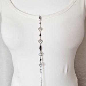 May include: A silver chain necklace with black beads and silver decorative charms. The necklace is displayed on a white tank top.