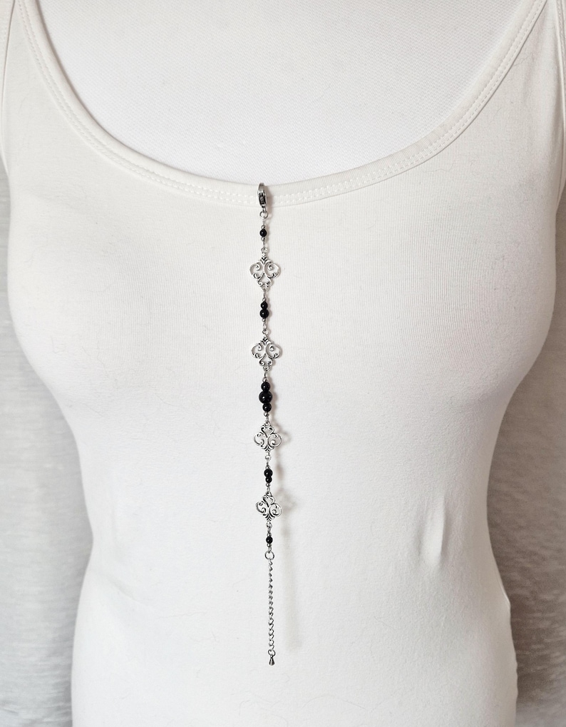 May include: A silver chain necklace with black beads and silver decorative charms. The necklace is displayed on a white tank top.