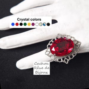 May include: A silver-toned ring featuring a large, oval-cut red crystal set in a decorative filigree frame. Above the ring, a color chart displays the text "Crystal colors" with nine circular swatches. Below, the text "Cachou Rêve de Bijoux" appears.