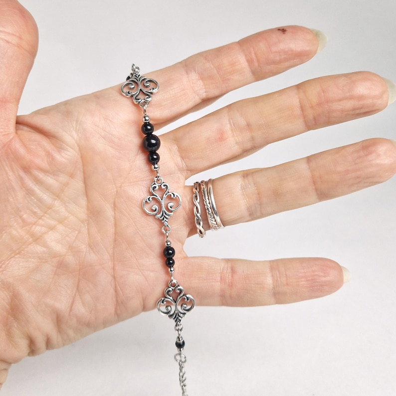 May include: A silver chain bracelet with black beads and ornate silver charms. The bracelet is on a person's hand.