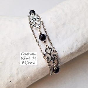 Peut inclure: Un bracelet en chaîne argentée avec des perles noires et des breloques argentées décoratives. Le bracelet est sur une surface blanche avec le texte "Cachou Rêve de Bijoux".