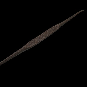 May include: A dark brown, elongated, pointed tool featuring a central section decorated with intricate, embossed floral and leaf patterns. The object tapers to thin, smooth points at both ends, with a textured surface throughout its length.