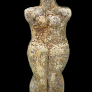 Peut inclure: Une petite figurine en pierre patinée avec une surface beige et brune marbrée. La sculpture présente une tête stylisée et allongée avec un nez proéminent, une poitrine définie et des hanches arrondies, se terminant par des jambes jointes.