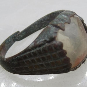 May include: A dark, weathered metal ring featuring a wide, textured band with a grid pattern and a central, smooth, translucent, off-white stone setting. The metal shows signs of oxidation and age, with a visible gap in the adjustable band.