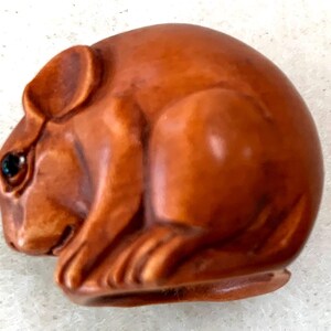 May include: A small, smooth, brown carved figurine of a crouching mouse with a rounded body, pointed ears, and a dark, inset eye. The sculpture features detailed textures on the face and paws, with a polished finish and a curved, compact posture.