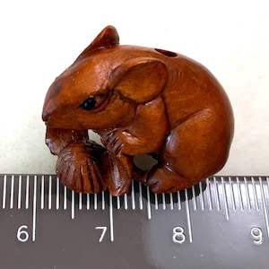 May include: A brown carved wooden figurine of a mouse holding a nut, positioned above a ruler showing a length of approximately 1.5 inches from the 6 to the 7.5 mark. The mouse has detailed facial features, small ears, and a smooth finish.