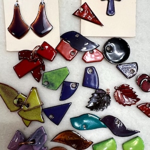 May include: A collection of assorted enameled metal jewelry components in various shapes and colors, including red, green, blue, purple, and brown. The set features geometric, leaf, and cross shapes, some with holes for hanging, alongside a copper penny.