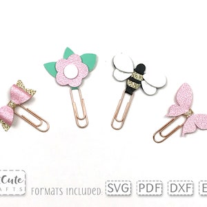 Op de afbeelding: Vier decoratieve paperclips met verschillende ontwerpen: een roze strik, een roze bloem met groene bladeren, een zwart-witte bij en een roze vlinder. De clips zijn goudkleurig.