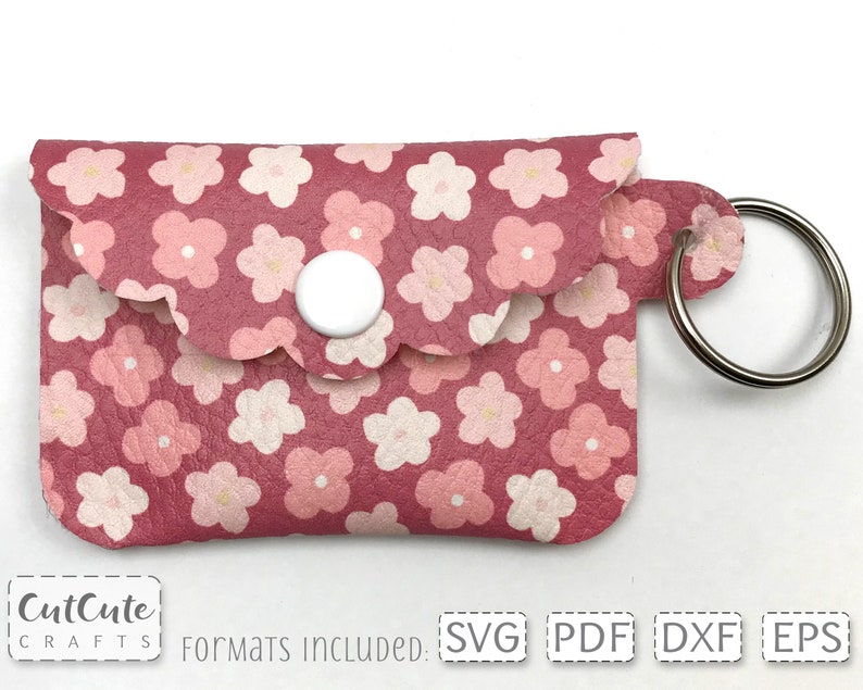 May include: A pink leather keychain with a floral pattern and a white button. The keychain has a scalloped edge and a silver key ring.