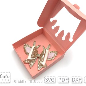 May include: A pink box with a cut-out crown design on the top. The box is open and a gold glitter bow with white and pink accents is inside. The text "Formats Included: SVG PDF DXF EPS" is at the bottom of the image.