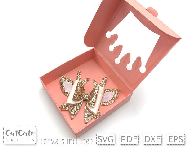 May include: A pink box with a cut-out crown design on the top. The box is open and a gold glitter bow with white and pink accents is inside. The text "Formats Included: SVG PDF DXF EPS" is at the bottom of the image.