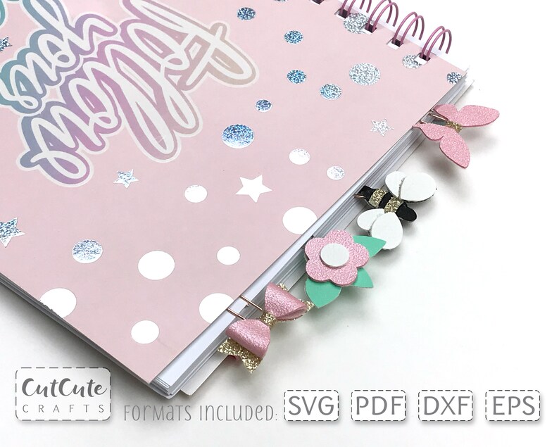 Op de afbeelding: Een roze notitieboek met een regenboog glitterontwerp en de woorden "Stay Happy" op de omslag. Het notitieboek heeft een spiraalbinding en is versierd met papieren uitsnijdingen van vlinders, bijen en bloemen.