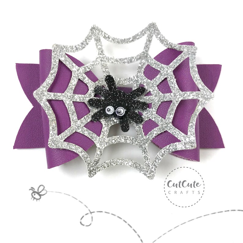 Op de afbeelding: Een paarse en zilveren glitter spinnenweb haar strik met een zwarte spin in het midden. De spin heeft knoopjesogen. De strik is handgemaakt en heeft een "CutCute Crafts" logo onderaan.
