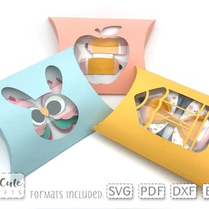 May include: Three pillow boxes with cut-out windows. The boxes are light blue, pink, and yellow. The windows show a pink and green owl, a red apple with a yellow pencil, and a yellow pencil with a blue bow tie.
