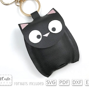 Könnte beinhalten: Schwarzer Leder-Schlüsselanhänger mit einem niedlichen Eulen-Design. Die Eule hat große weiße Augen, eine rosa Nase und rosa Ohren. Der Schlüsselanhänger hat einen goldenen Metallring.