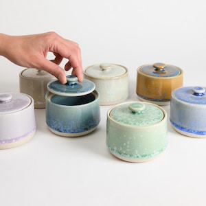 May include: A selection of ceramic lidded jars in shades of blue, green, and yellow. The jars feature a speckled glaze and a circular form. One jar's lid is being lifted by a hand.