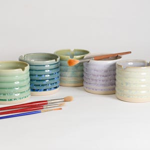 May include: Handmade ceramic ashtrays in shades of green, blue, and lavender. Each ashtray has a ribbed design and a small notch for cigarettes. Paintbrushes are also present, suggesting an artistic theme. Measurements are in centimetres.