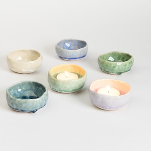 May include: A selection of small, handmade ceramic bowls in various colours, including cream, blue, and green. Some bowls contain lit tea light candles. The bowls have a textured, organic shape, ideal for home decor.