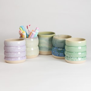 May include: A selection of handmade ceramic pen holders in various colours and designs. The holders are cylindrical, with some featuring ribbed textures and others with unique patterns. Some holders contain colourful pens and markers.
