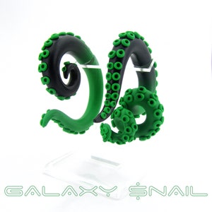 May include: A pair of black and green octopus tentacle-shaped ear gauges. The gauges are made of polymer clay and are designed to look like octopus tentacles. The gauges are on a clear display stand.