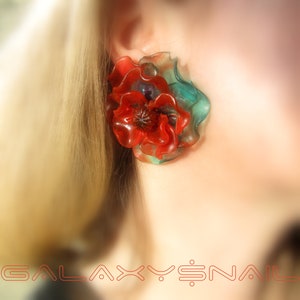 Puede incluir: Pendientes de flor de resina roja y verde. La flor es una amapola estilizada con un diseño en capas. Los pendientes son una pieza llamativa y serían una gran adición a cualquier colección de joyas.