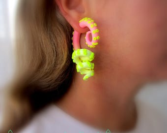 May include: Pink and neon yellow octopus tentacle earrings. The earrings are worn in the ear and feature a spiral design with small, round details. The earrings are a vibrant accessory.