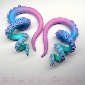 May include: A pair of galaxy-themed octopus ear gauges. The gauges are made of polymer clay and feature a gradient of pink and blue colors. The octopus tentacles are curled and have purple accents.